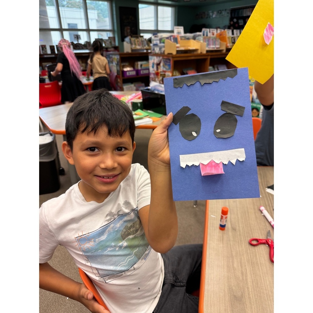 A child proudly holds up a blue monster face craft with pink tongue.