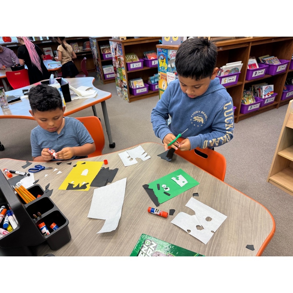 Two children work at a table with scissors and glue, making colorful monster faces.