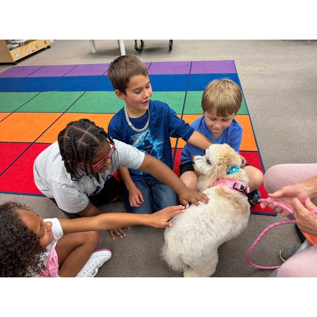 A group of children sit on a colorful carpet, smiling and petting a small white therapy dog.