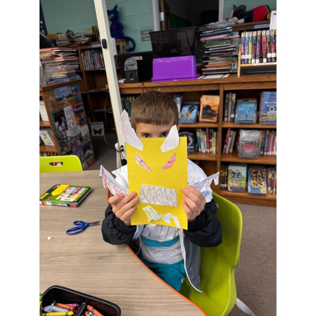 A child holds up a yellow paper with sharp teeth and hand-drawn red eyes