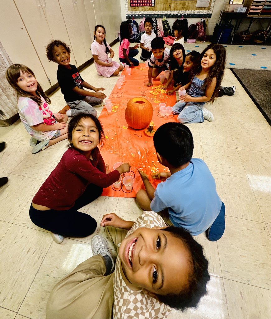 Smiling students sitting around the pumpkin seed activity, with some holding cups and others looking toward the camera.
