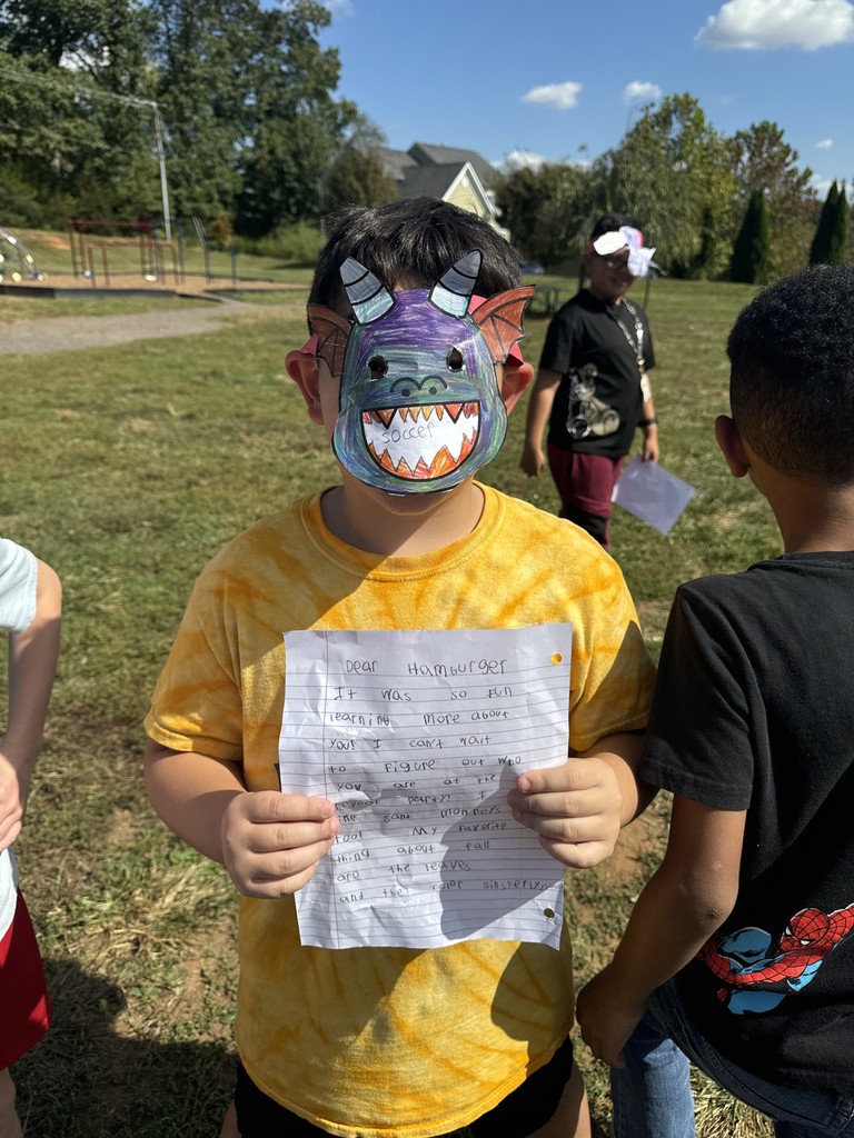 student wearing a yellow tie-dye shirt and a handmade purple and green dragon mask holds up a handwritten letter. Other students are nearby on the playground enjoying the sunny day.