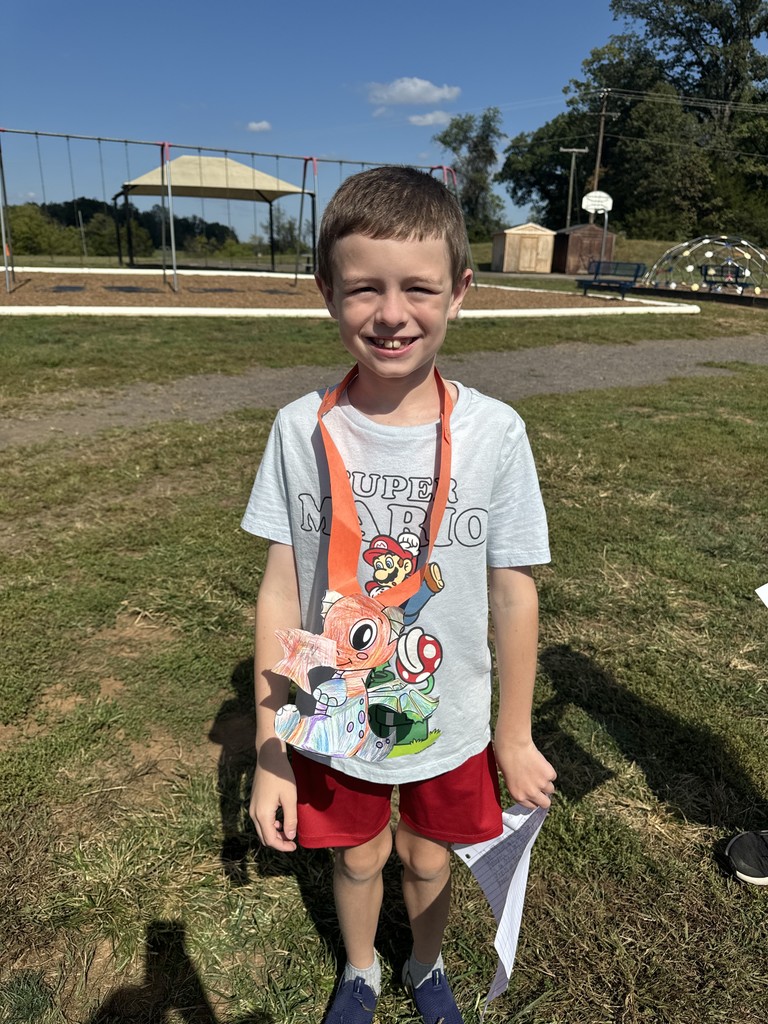 A smiling student wearing a gray Super Mario shirt and red shorts stands on the playground holding a colorful dragon craft. The swing set and climbing dome are visible in the background under a clear, sunny sky.