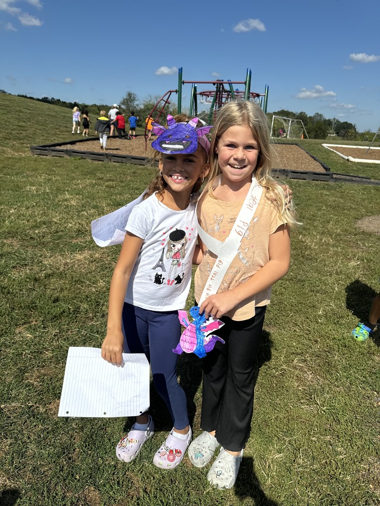 Two smiling students stand side by side on the playground. One is wearing a purple dragon mask with wings and holding a sheet of paper, while the other wears a paper sash and holds a colorful dragon craft. Children can be seen playing and walking in the background under a bright blue sky.