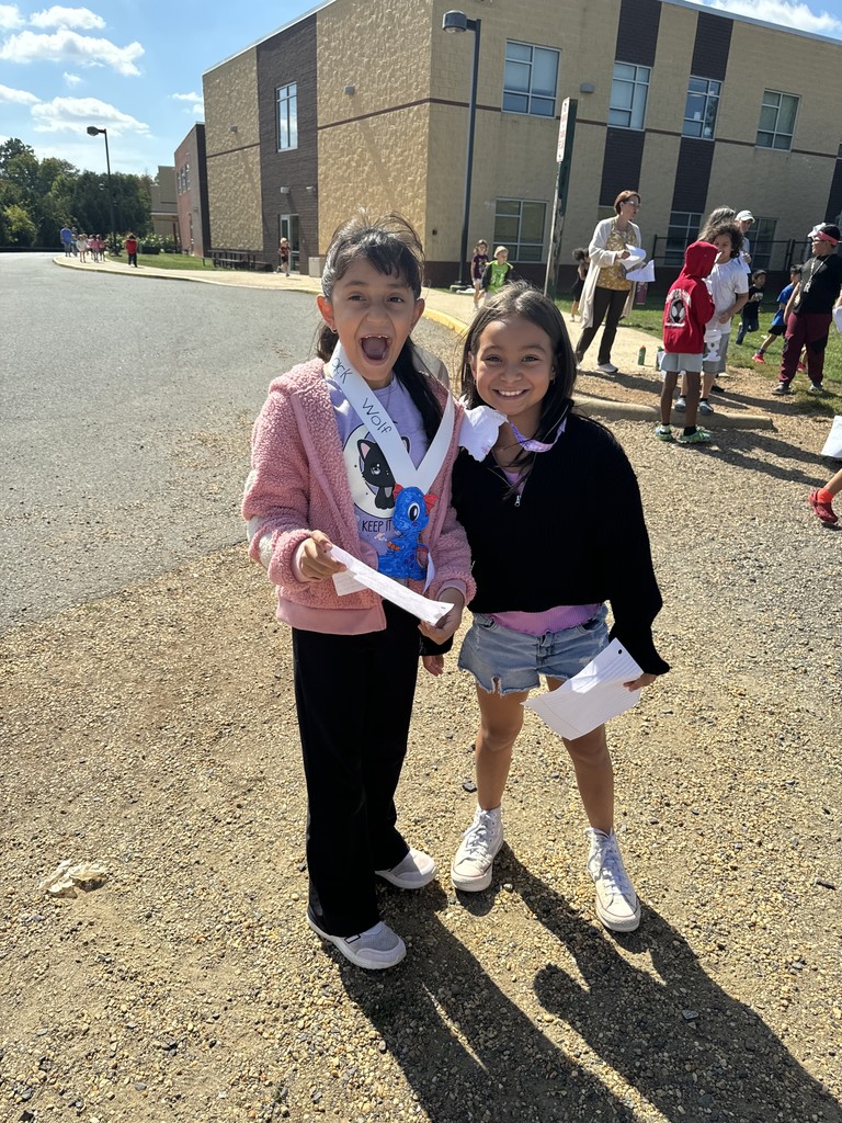 Two students smile and pose together outside near the school building. One holds a blue dragon craft and wears a paper sash while the other stands beside her with a paper in hand. Other students and teachers are visible in the background enjoying the outdoor activity.
