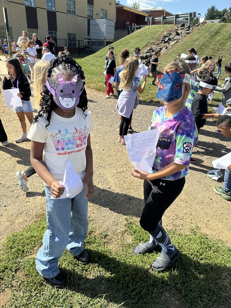 Two students wearing handmade dragon masks stand outdoors holding papers. One wears a white Hello Kitty shirt, and the other wears a tie-dye shirt and boots. A group of classmates is gathered behind them near a grassy hill.