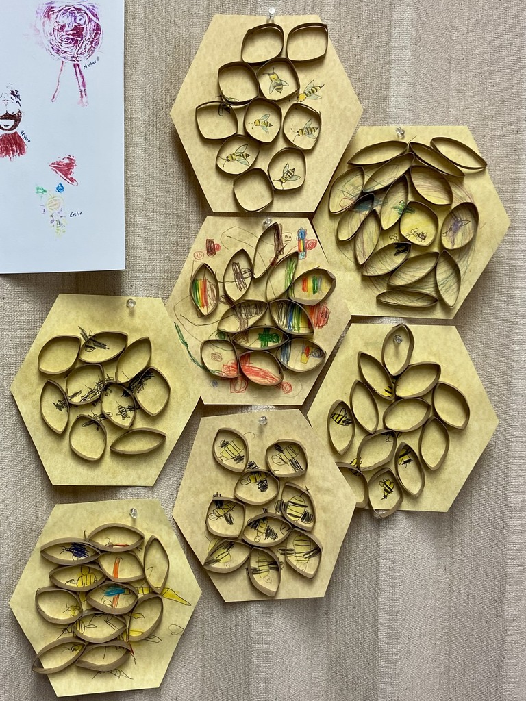 A wall display features several yellow hexagon-shaped pieces of paper arranged like a honeycomb. Each hexagon has oval shapes made from rolled brown paper forming cells, with drawings of bees and colorful artwork inside. A student-drawn poster with portraits and names is pinned above the honeycomb display.