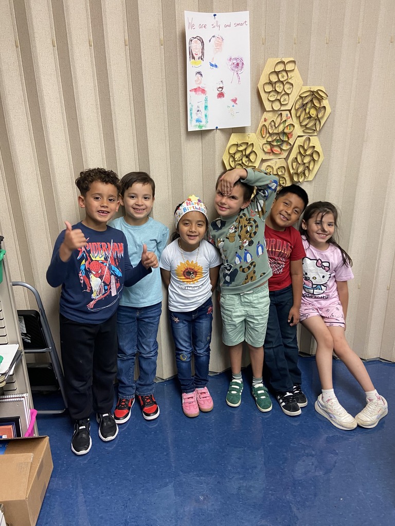 Six young students pose proudly in front of the classroom’s honeycomb art display. Above the display is a student-drawn poster that reads “We are silly and smart.” The children smile and give thumbs up, showing enthusiasm for their project.