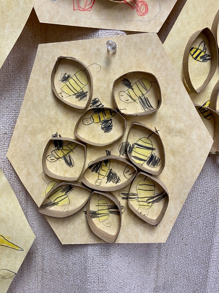 A close-up of one hexagon from the honeycomb art display shows several small paper ovals glued to the hexagon base. Each oval contains a child’s drawing of a bee with a yellow body and black stripes.