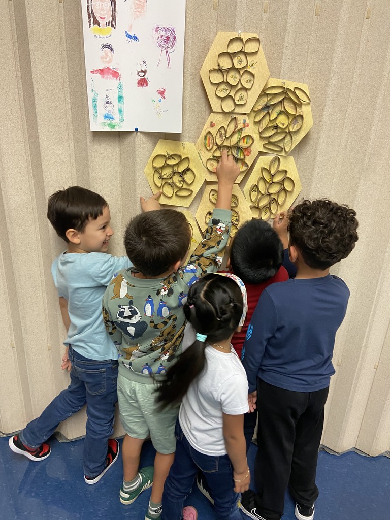 Five young students stand close together, pointing excitedly at the honeycomb art display on the wall. They are smiling and engaged while observing the bee drawings and colorful designs within the hexagons.