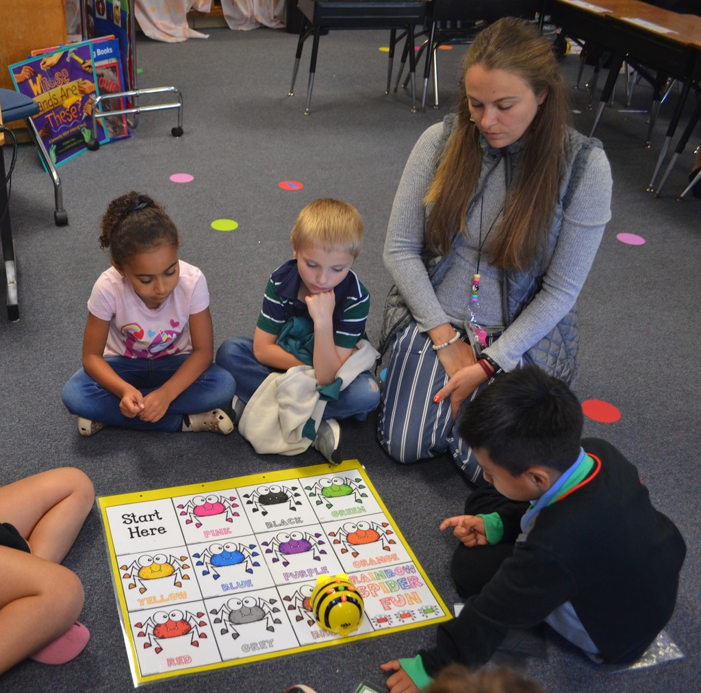 3 students and their teacher work on the classroom floor, using a Beebot
