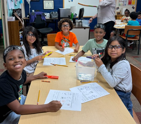 Five students sit at a table wearing safety goggles and holding pencils. A small plastic container and science worksheets are on the table, and all are smiling at the camera.