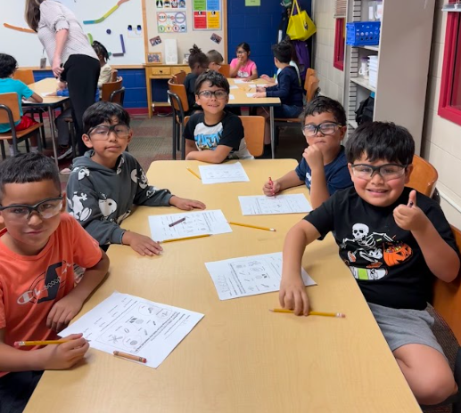 Five smiling students wearing safety goggles sit around a classroom table with science worksheets and pencils in front of them. One student gives a thumbs-up gesture toward the camera.