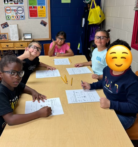 Five students sit together at a rectangular classroom table, smiling and holding pencils while working on science worksheets. They are wearing protective safety goggles and looking toward the camera.