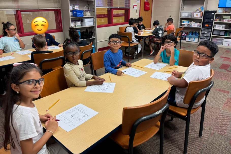Six students sit at a classroom table wearing safety goggles and holding pencils while completing science worksheets. Other groups of students can be seen working at tables in the background.