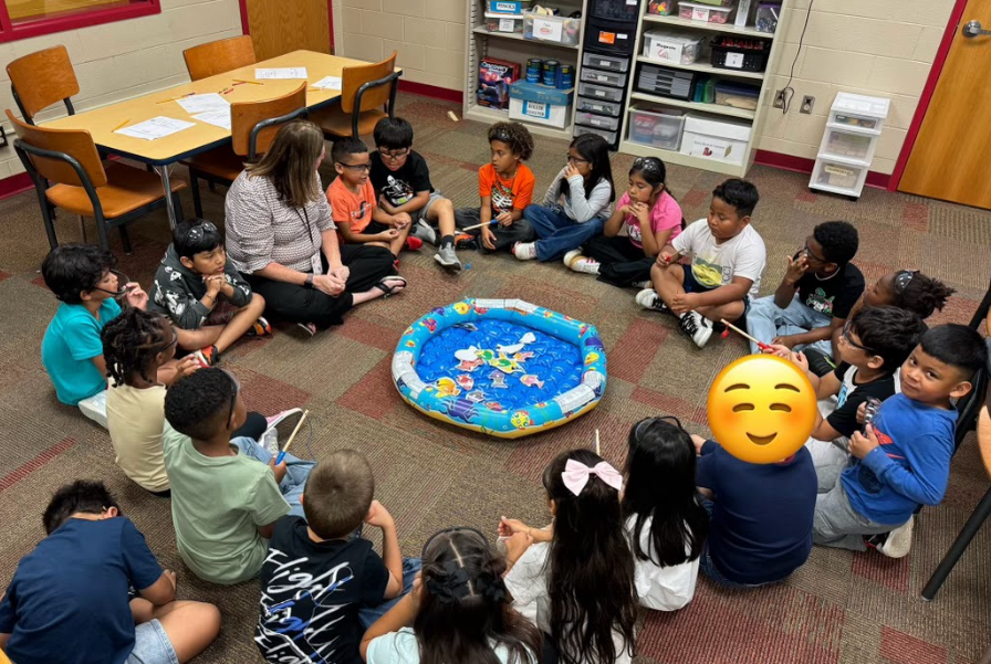 A large group of students sits in a circle on the classroom floor around a small inflatable pool filled with colorful paper fish. A teacher sits with them, and students hold magnetic fishing rods as part of a science activity.