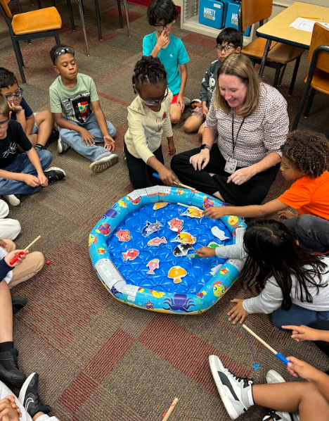 Students sit on the classroom floor in a circle around an inflatable pool filled with colorful fish cutouts. A teacher kneels beside them, smiling, as students use fishing rods to “catch” the paper fish during a hands-on activity.