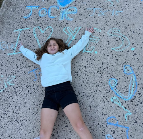 A student lies on the sidewalk with arms spread out, surrounded by blue chalk drawings and words that include “I ❤️ YES” and her name written nearby.