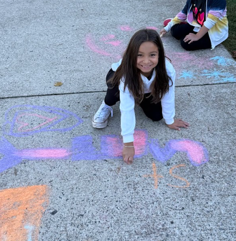 A student kneels on the sidewalk, smiling as she adds to a large colorful chalk design that includes the word “Yowell” and various shapes and hearts.