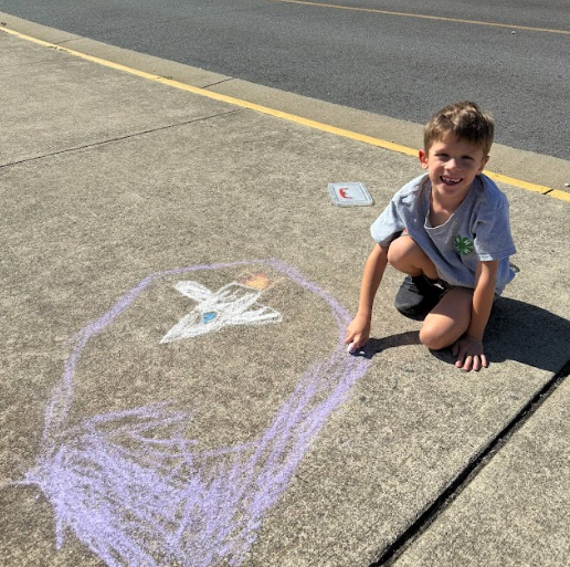 A young student kneels on the sidewalk next to a purple chalk drawing of a rocket ship with a small flame at the bottom, smiling at the camera.
