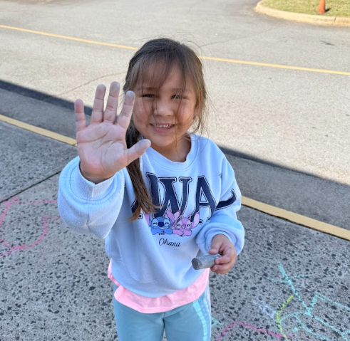 A smiling student wearing a light blue “Ohana” sweatshirt raises a chalk-covered hand toward the camera while holding a piece of sidewalk chalk in the other hand.