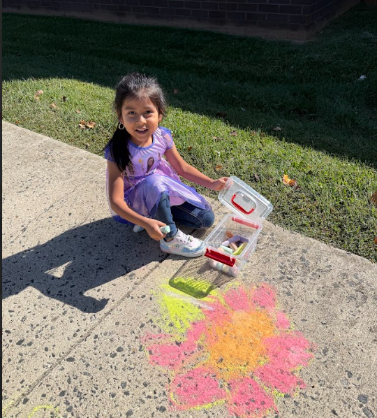 A student wearing a purple dress sits on the sidewalk next to her chalk artwork—a large orange and pink flower with green leaves—while holding a piece of chalk and smiling.