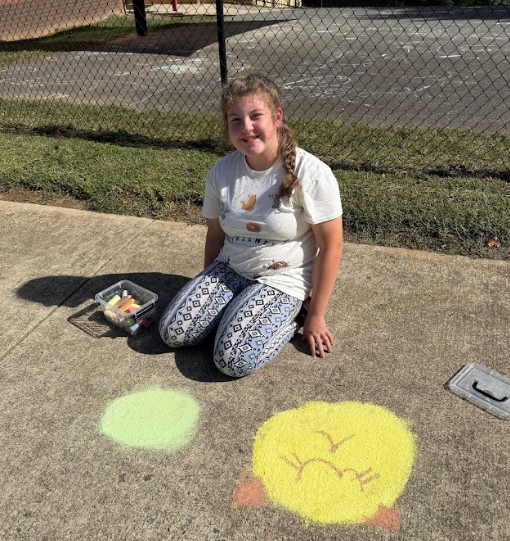 A student sits on the sidewalk with a small container of chalk beside her, smiling as she creates colorful chalk art that includes a bright yellow circle and a green circle.