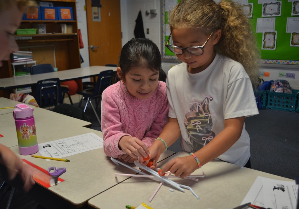 A group of students is working to add candy pumpkins to a straw STEAM design on their desks in their classroom.