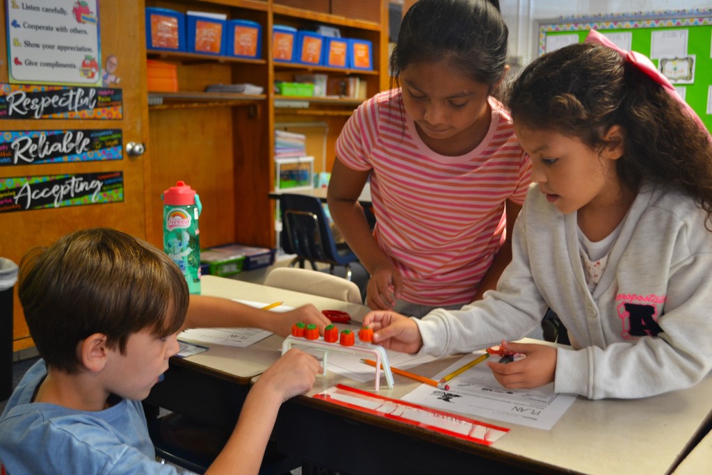 A group of 3 students is working to add candy pumpkins to a straw STEAM design on top of their desks. Respectful, Reliable, Accepting in on a poster in the back.