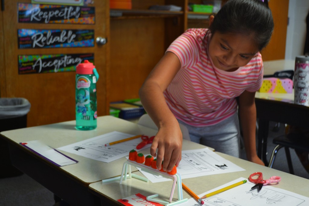 Girl student adds a candy pumpkin to a straw bridge design on a desk.  Water bottle in the background.  Respectful, Reliable, Accepting in on a poster in the back.