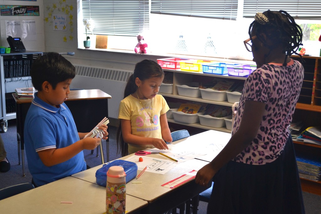 A group of students are working their materials (straw, tape, pencil) to build a design at their desks in a school classroom.