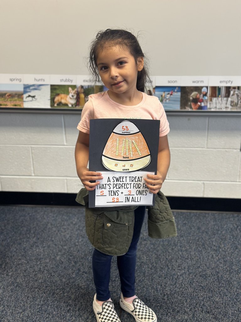 Student proudly displaying her candy-corn math craft — Her poster shows “53 in all,” neatly decorated and held up in front of her, as she smiles for the photo.