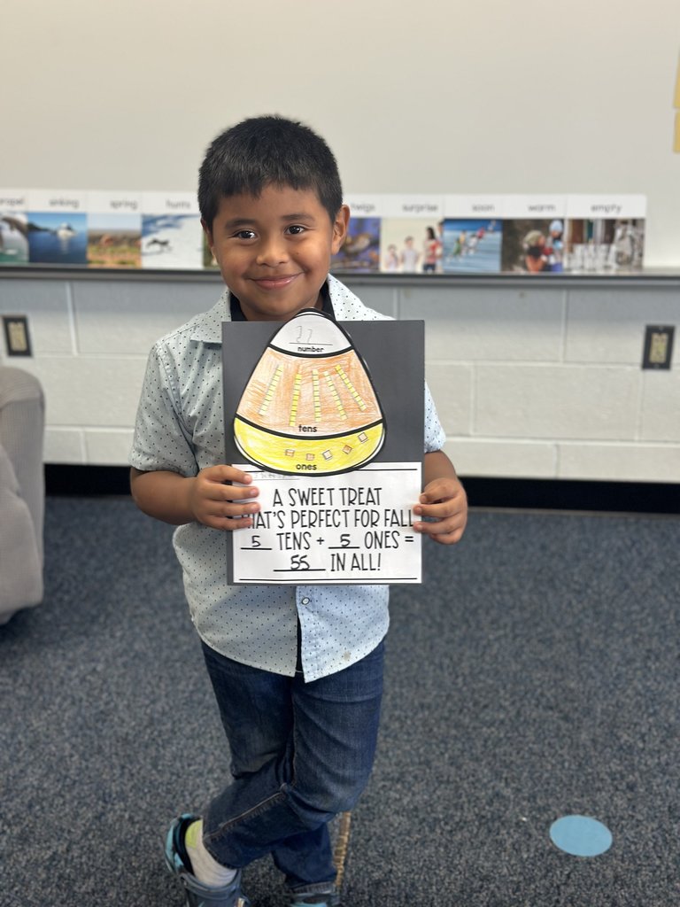 Smiling student holding finished candy-corn place value project — His poster shows “55 in all” with five tens and five ones, colored brightly in orange and yellow.