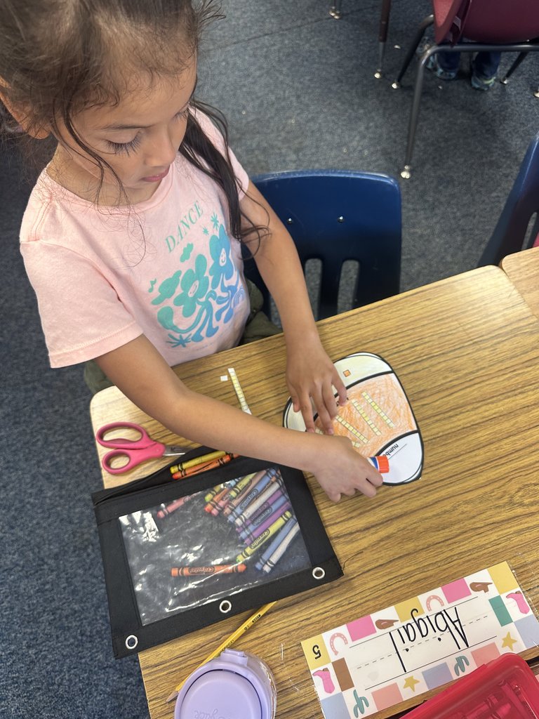 Student gluing pieces to complete a candy-corn math craft — She’s focused on carefully attaching place-value blocks, with a colorful crayon pouch open beside her.