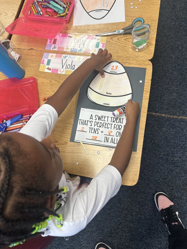 Girl assembling a candy-corn place value activity — She’s gluing strips to represent tens and ones, working on a fall-themed math project with her name tag and bright supplies on the desk.