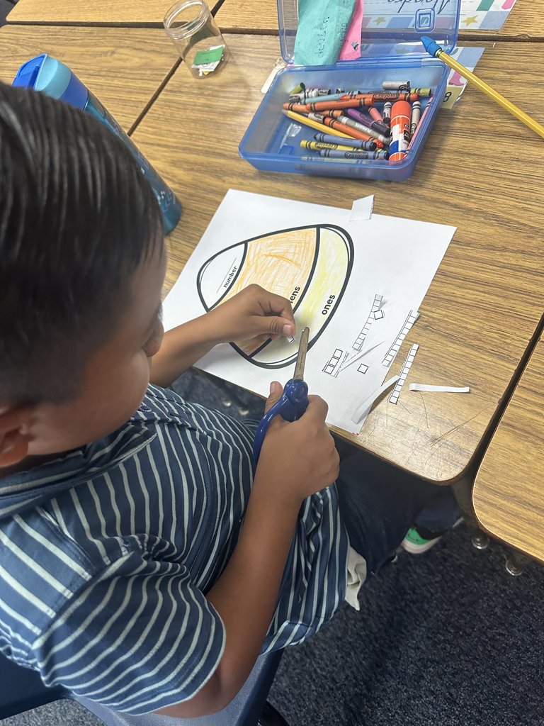 Boy cutting out a candy-corn shaped math craft — He’s seated at his desk, using scissors to trim a paper divided into “hundreds, tens, and ones” sections, with crayons and supplies spread out beside him.