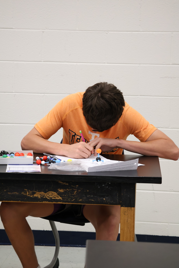 A student in an orange shirt leans over his work at a lab table, assembling molecular models and writing on a worksheet. Small colorful model pieces are scattered on the table beside him.