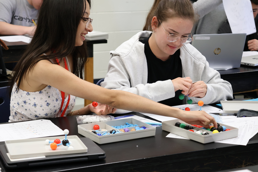 Two students smile as they work together at a lab table, building molecular models with colorful plastic atoms and connectors. Papers, trays of model pieces, and laptops are spread across the table.
