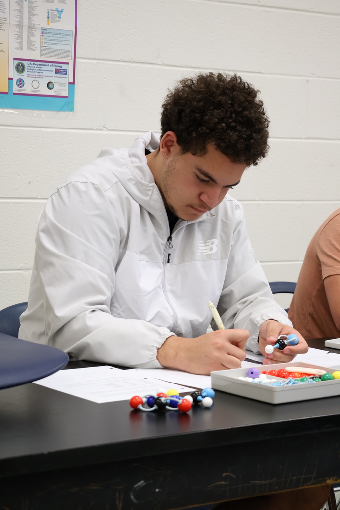 A student in a white jacket concentrates on building a molecular model while holding a pencil. Trays of model parts and a completed molecule rest on the desk in front of him.