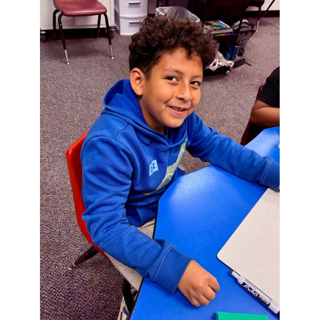 A student in a blue hoodie smiles at the camera while sitting at a blue table with math manipulatives
