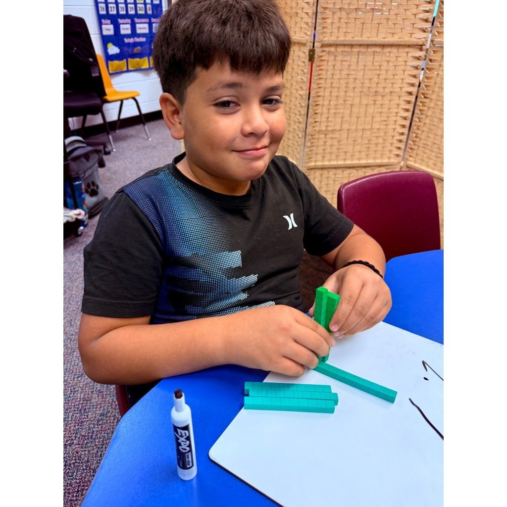 A student in a black shirt works with base-ten blocks on a whiteboard, smiling slightly.