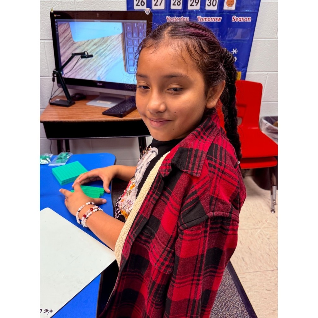 A student with braids and a red plaid shirt sits at a blue table, holding base-ten blocks with a computer in the background.