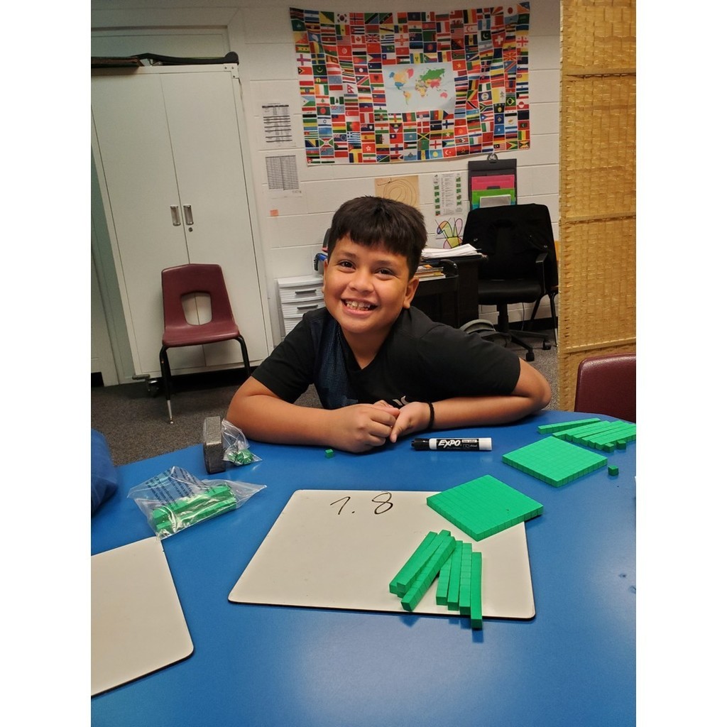 A smiling student sits at a blue table with base-ten math blocks and a whiteboard showing the number 1.8.