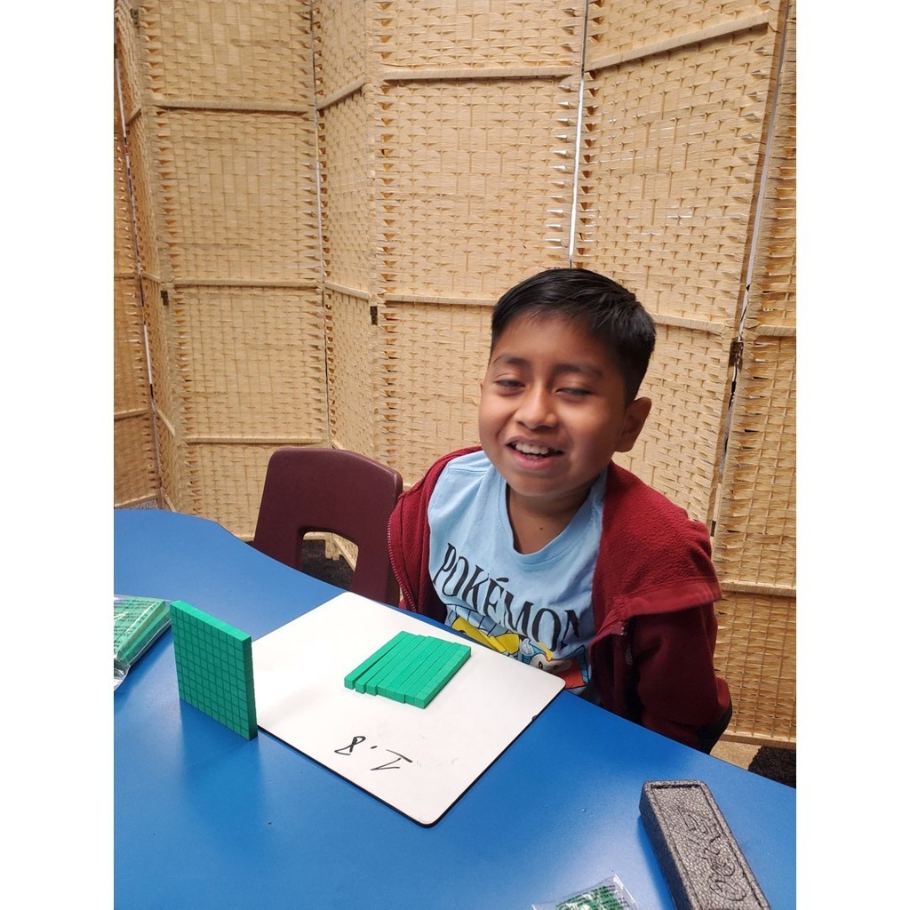 A student in a red jacket smiles while working with green base-ten blocks on a whiteboard showing the number 8.7.