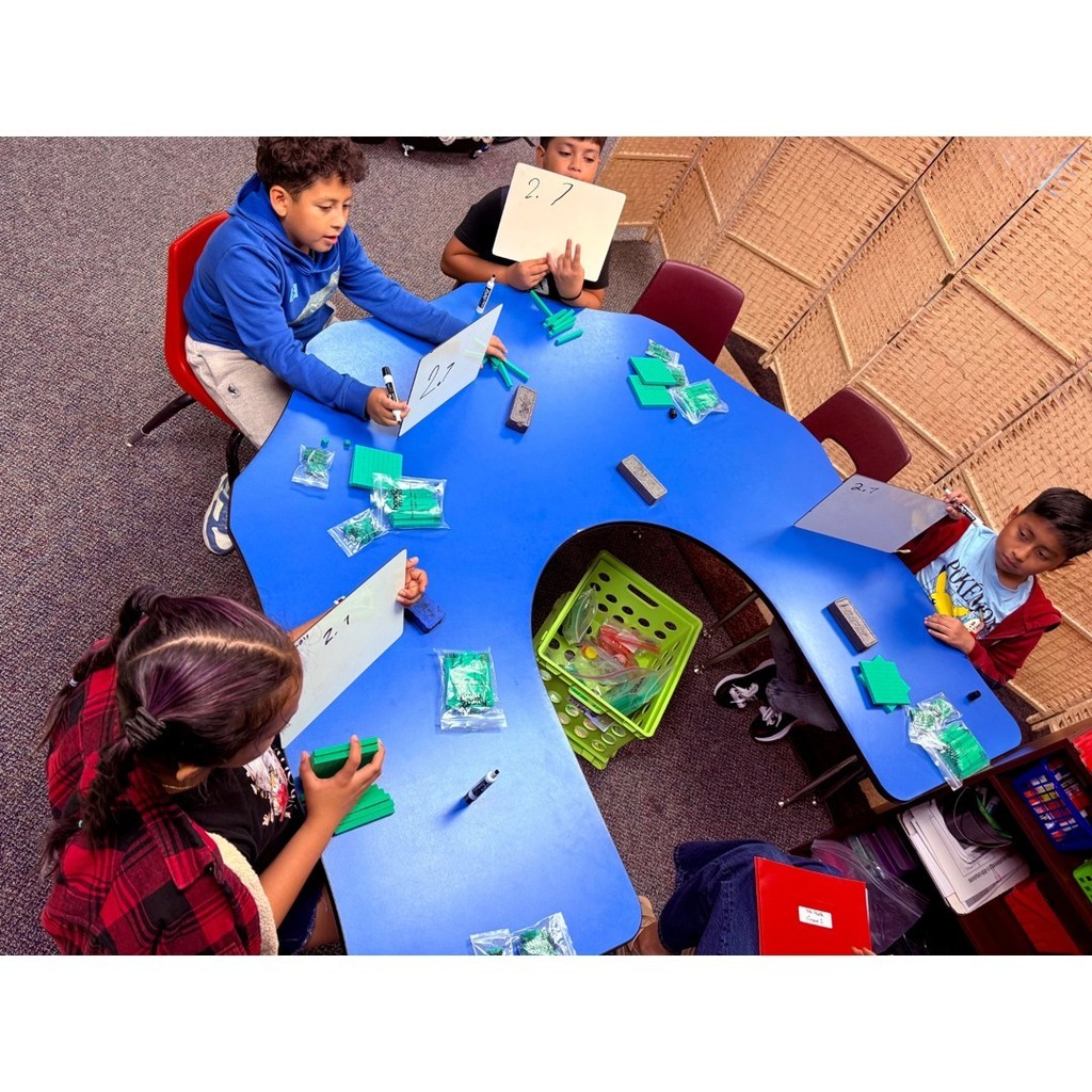 Four students sit around a blue horseshoe table, each holding whiteboards with decimal numbers and using base-ten blocks.