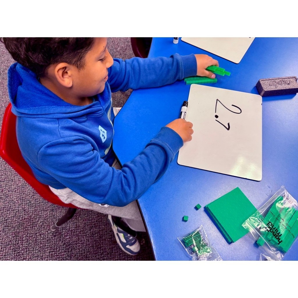 A student in a blue hoodie arranges base-ten blocks on a table while writing 2.7 on a whiteboard.