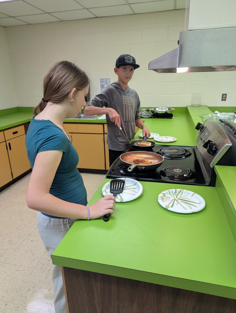 Two young people, a girl and a boy, cooking pancakes on an electric stovetop in a commercial or classroom kitchen. The girl on the left, wearing a teal top, holds a spatula over an empty plate, looking at the pan. The boy behind her, wearing a baseball cap, uses a spatula to flip a pancake in a second pan. The kitchen has distinctive bright green countertops and dark wood cabinetry.