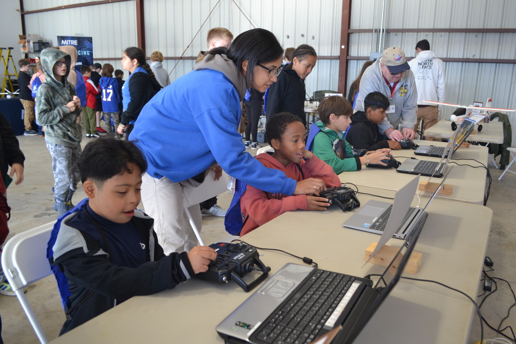 Image contains a large group of students in an airport hanger.  At the station are computers on tables.  Each computer has a student working and adults helping.  Additional students in the background.