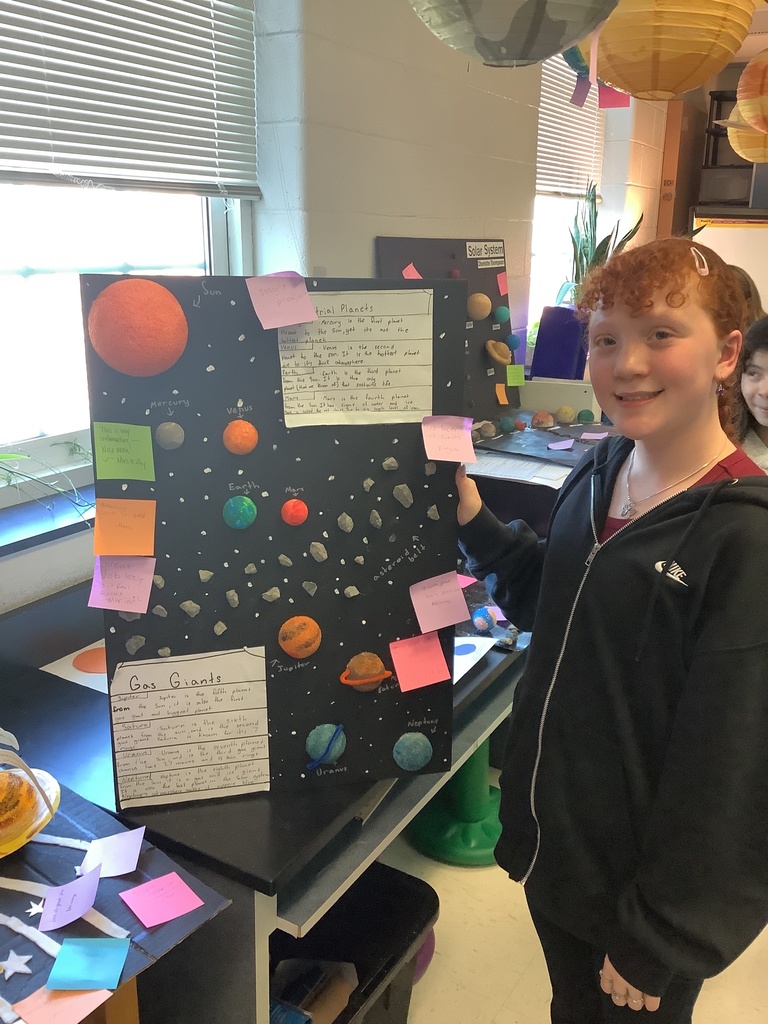 A student presents a colorful solar system poster with labeled planets and notes in a classroom setting. They smile proudly, conveying excitement and achievement.