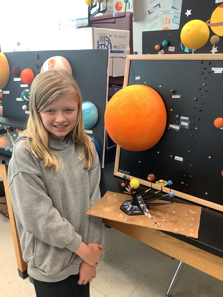 A smiling child in a gray hoodie stands next to a solar system display. Large colorful planets are mounted on black boards in a classroom setting.